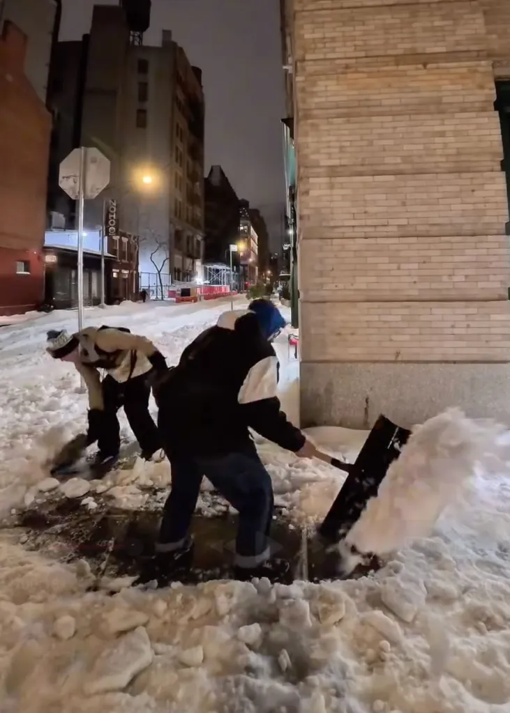 Two people shovel snow on a city street at night.